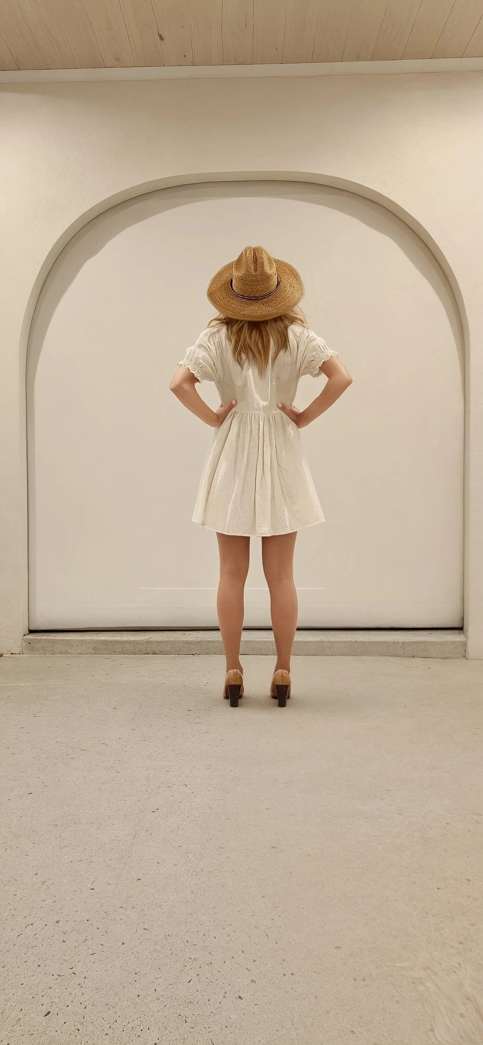Back view of model with hands on hips, wearing Auguste The Label Mara Mini dress worn with a straw hat and tan heels, styled in a relaxed neutral space.