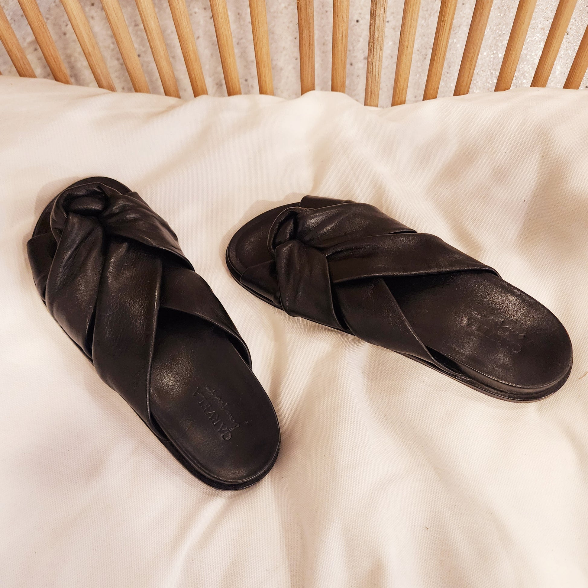 Carvela black leather slides styled on cream cotton with timber backdrop, highlighting laid-back luxe appeal