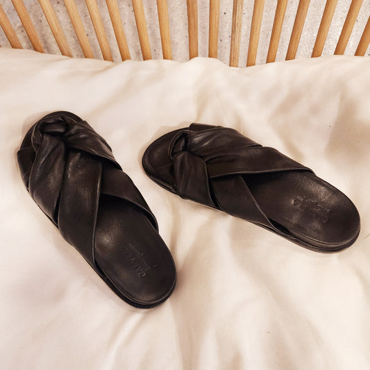Carvela black leather slides styled on cream cotton with timber backdrop, highlighting laid-back luxe appeal