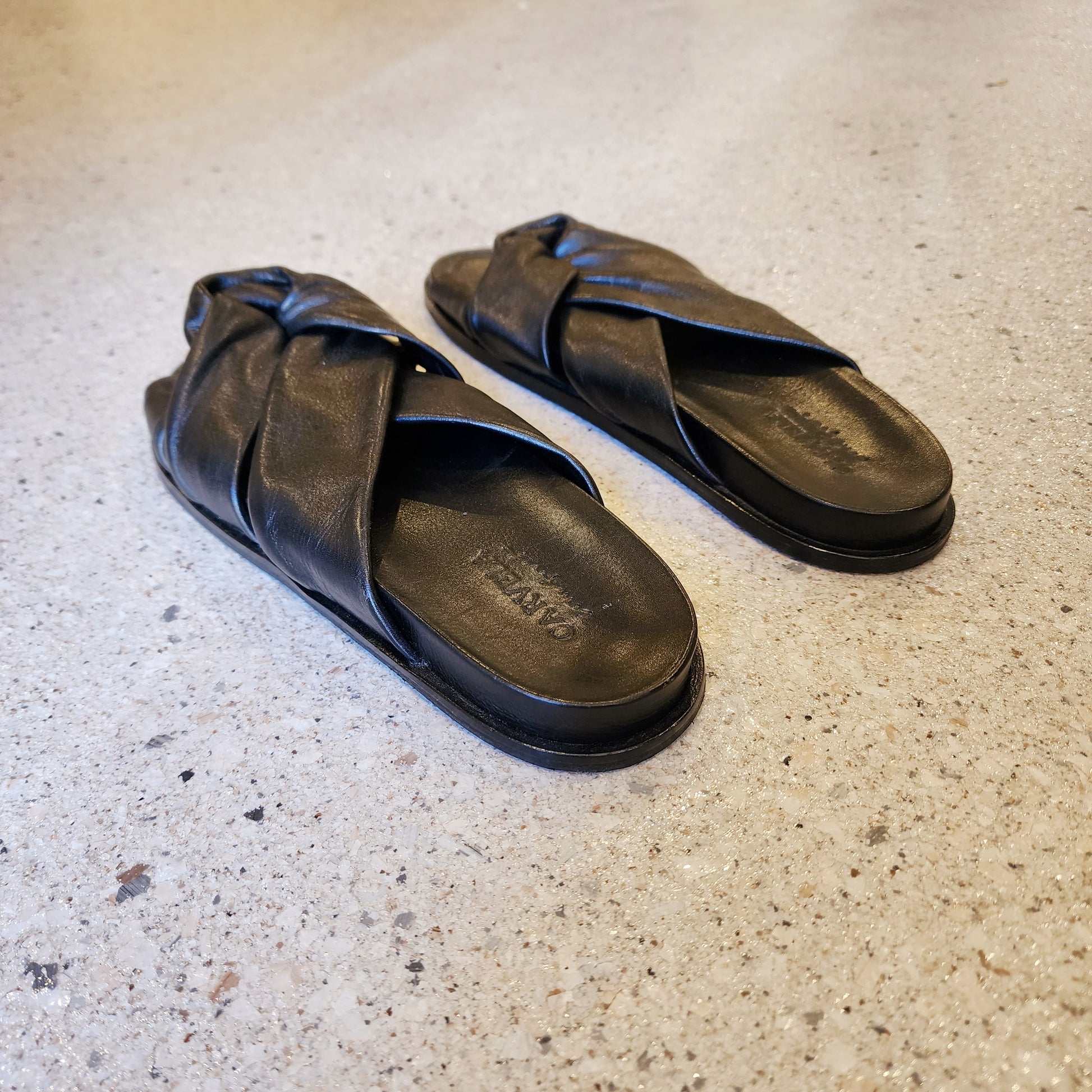 Carvela black leather knot slides photographed on terrazzo-style concrete flooring, showing back and top cross-strap detail