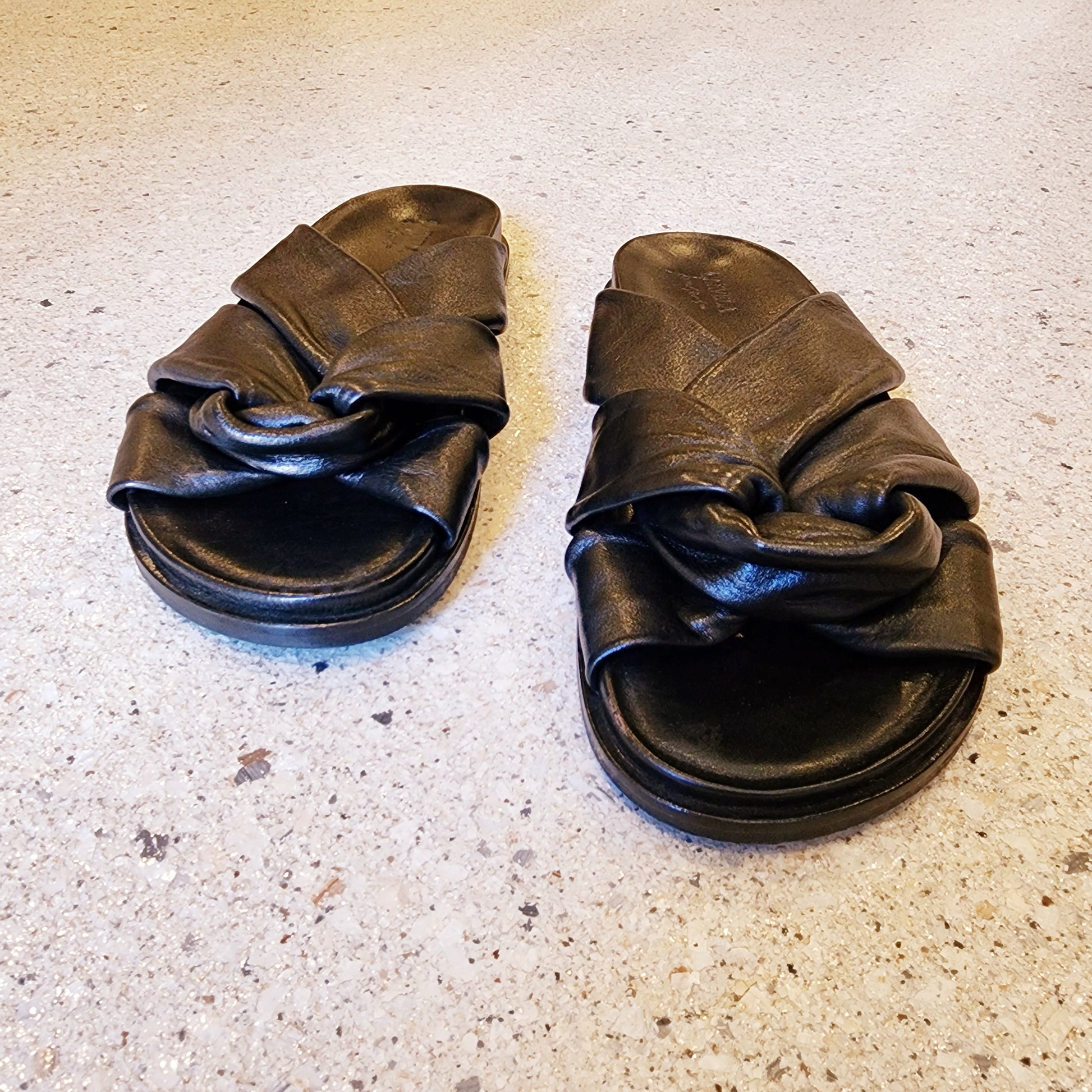 Carvela black leather knot slides photographed on terrazzo-style concrete flooring, showing front and top cross-strap detail.