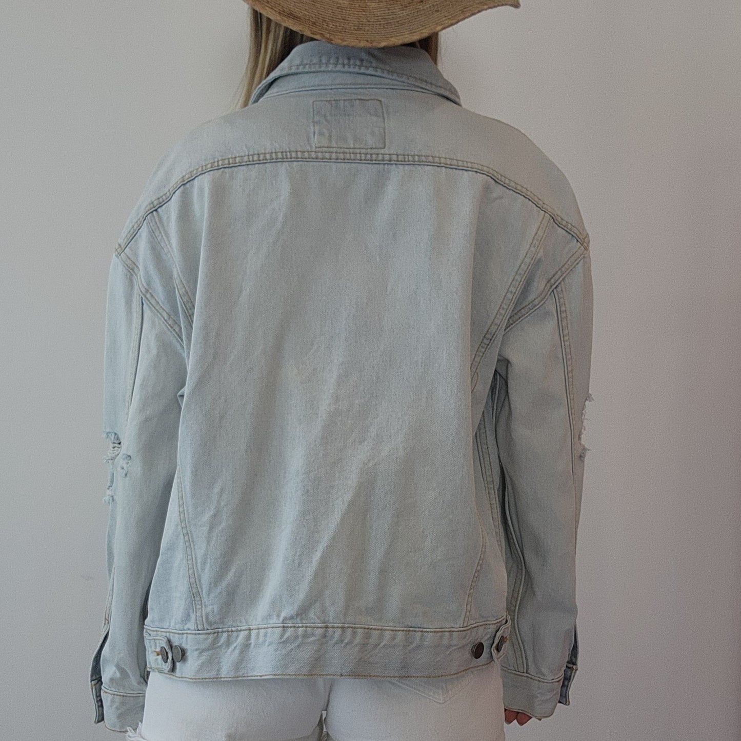 Back view of a model wearing a light wash denim button up jacket by clothing designer RVCA. Distressed style, made to last.