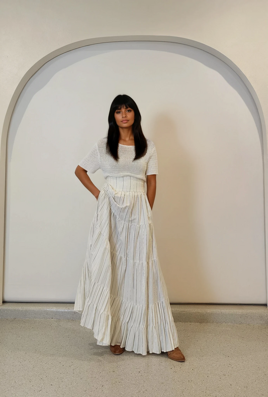 Model wearing off-white short-sleeve knit top, paired with a striped maxi skirt. The top is form-fitting with a slightly open weave and classic crew neckline.
