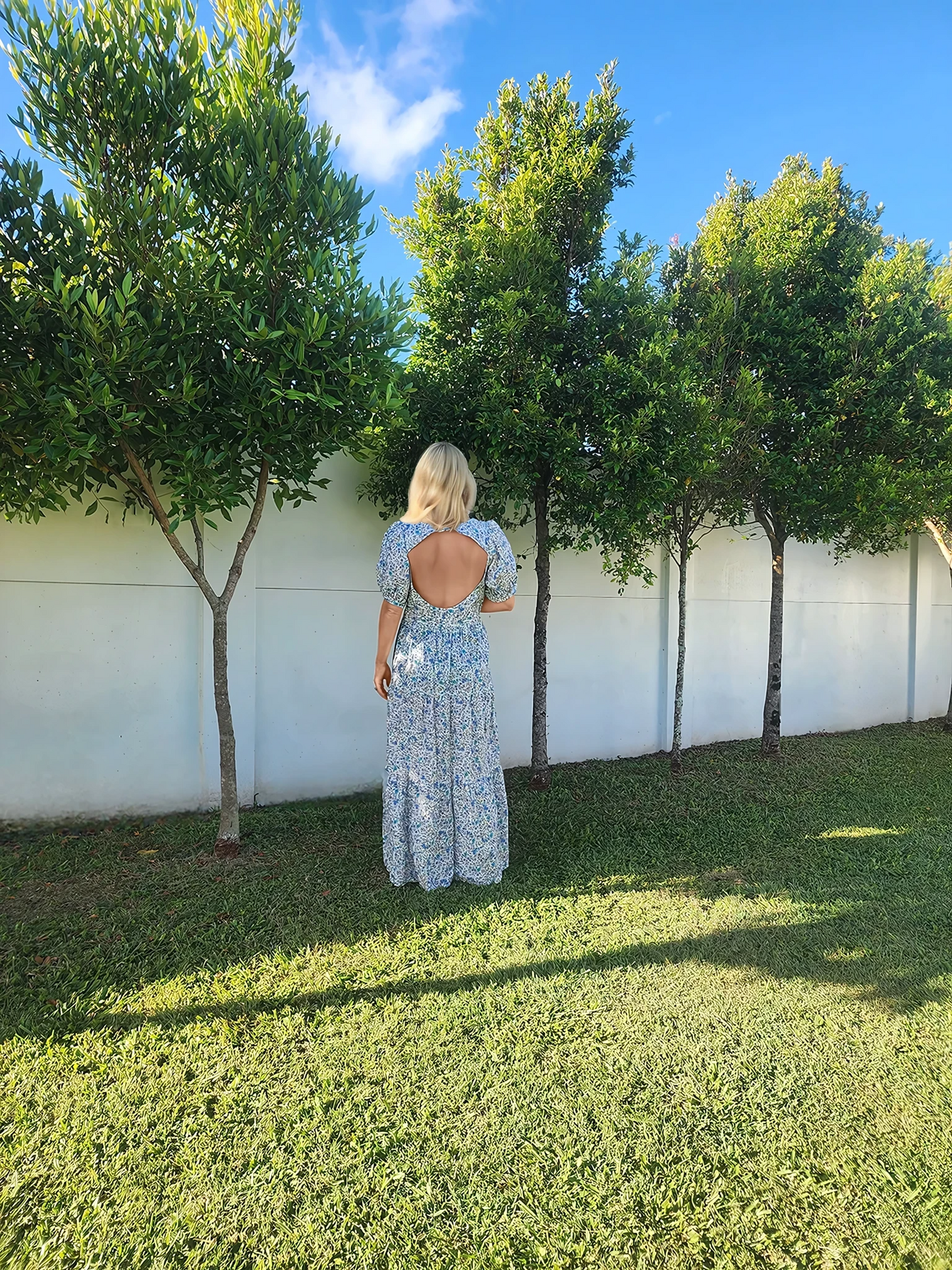 Model showing the back view of Oak Meadow blue floral maxi dress, with an open back and puff sleeves.