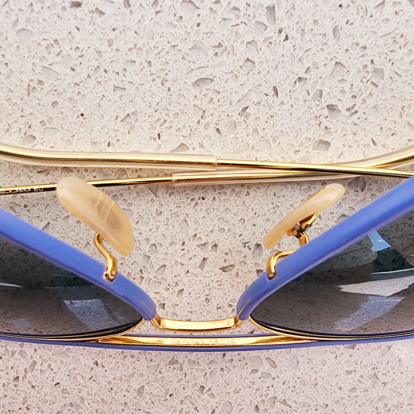 Close-up of Ray Ban aviator Sunglasses with blue lenses, gold frames, and beige nose pieces on a textured surface.
