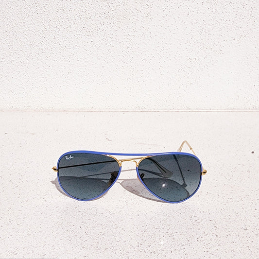 Ray Ban full colour aviator sunglasses with blue lenses and gold frame on a white textured surface.