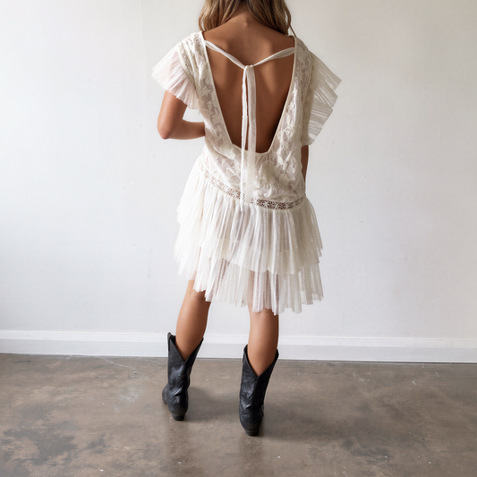 Back view of woman wearing a short white lace dress with a deep open back and ruffled tulle hem, paired with black cowboy boots.
