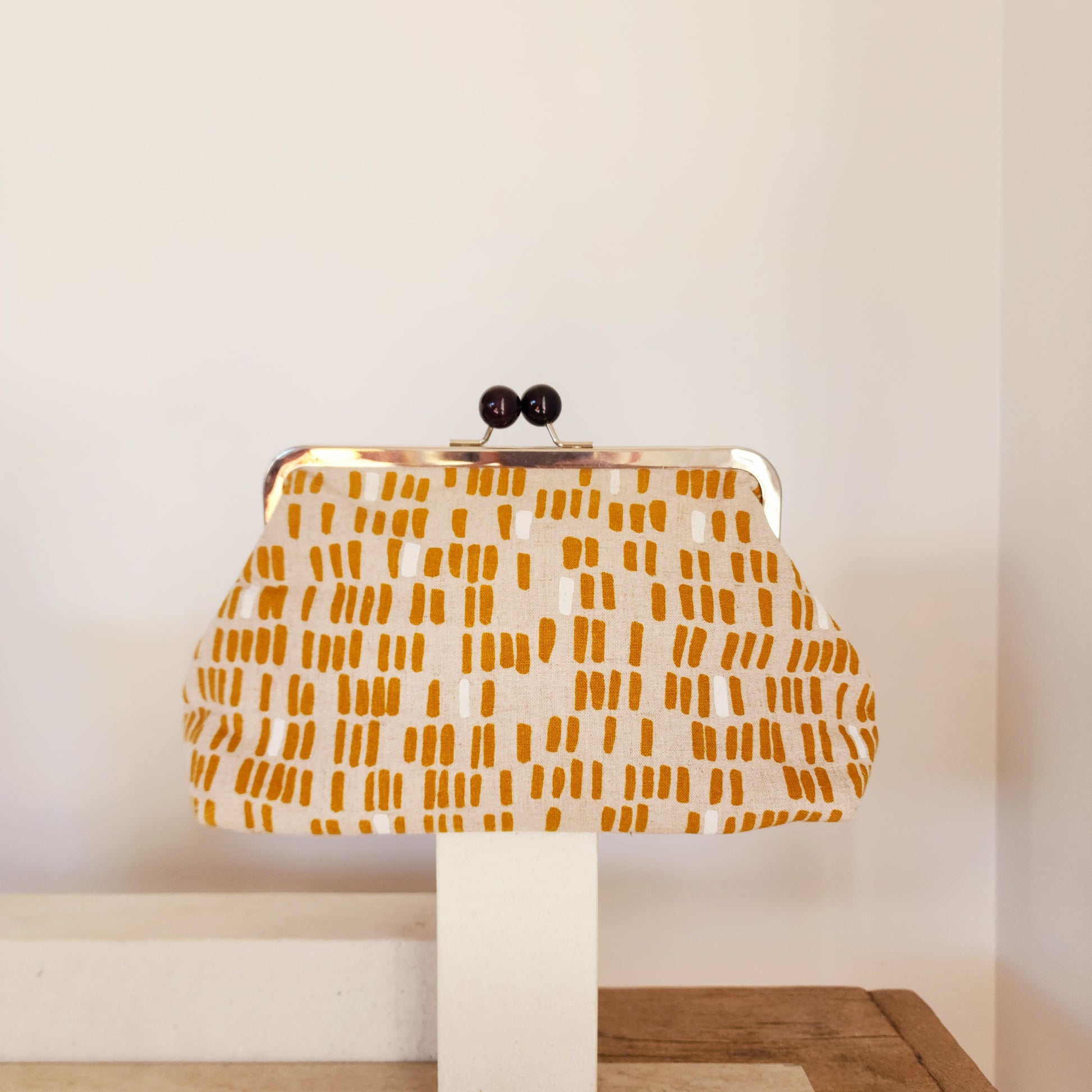 A retro clutch bag with a mustard line design displayed on a stand against a neutral background.