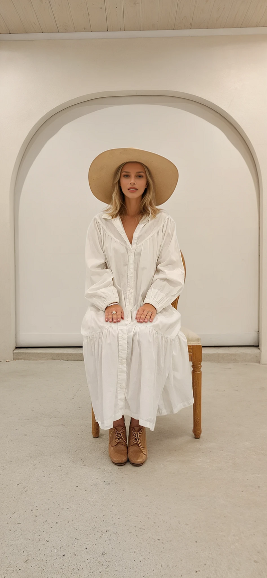 Model seated front-facing in white maxi dress and wide-brimmed hat, hands on knees