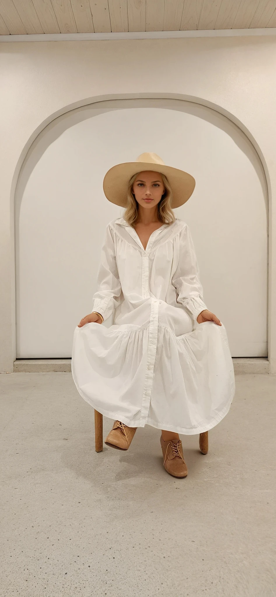 Model seated, holding out skirt of white maxi dress to show volume and gathered detail
