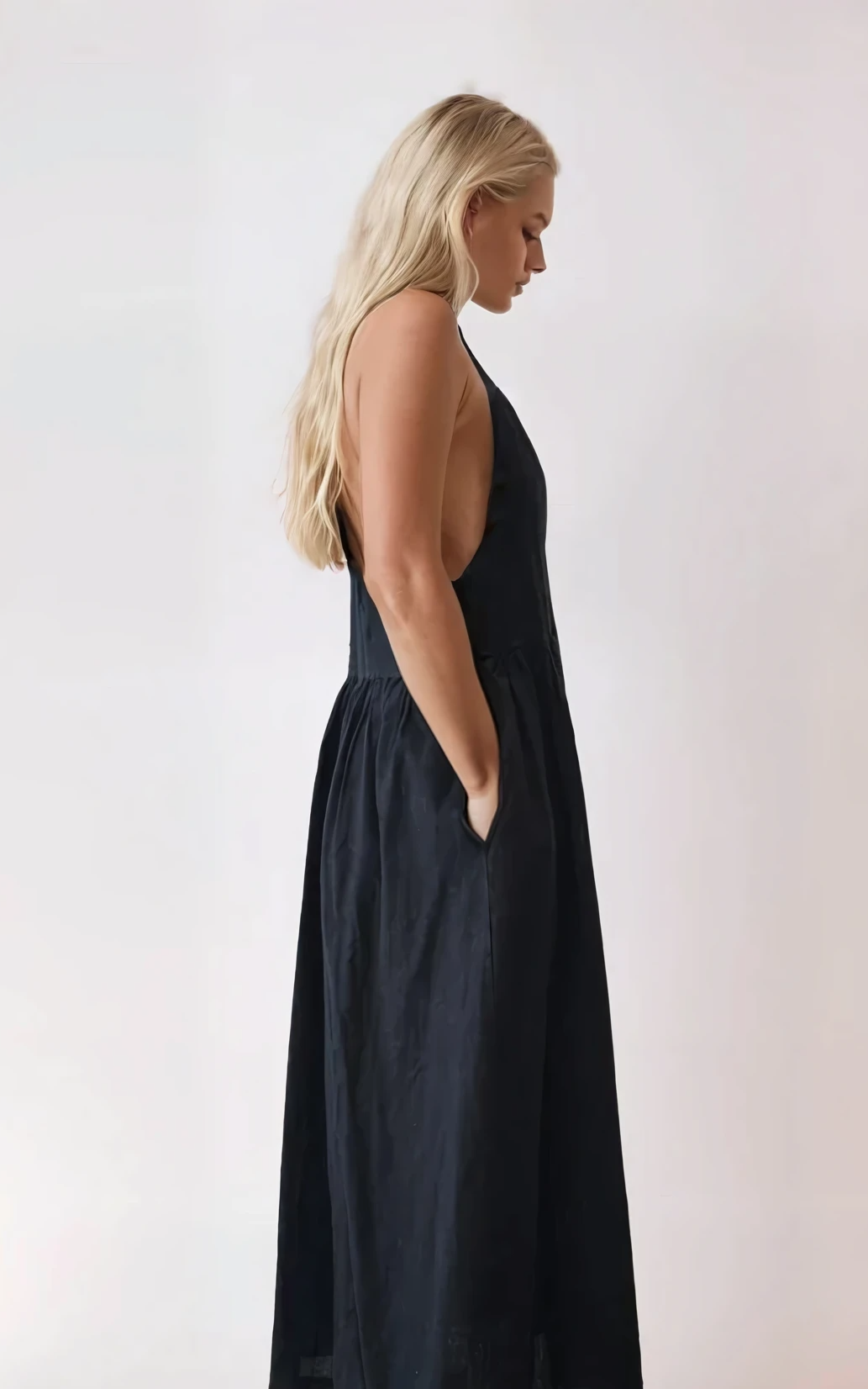 Side profile of black pinafore dress showing open sides, deep armholes, and relaxed fit with hands in pockets.