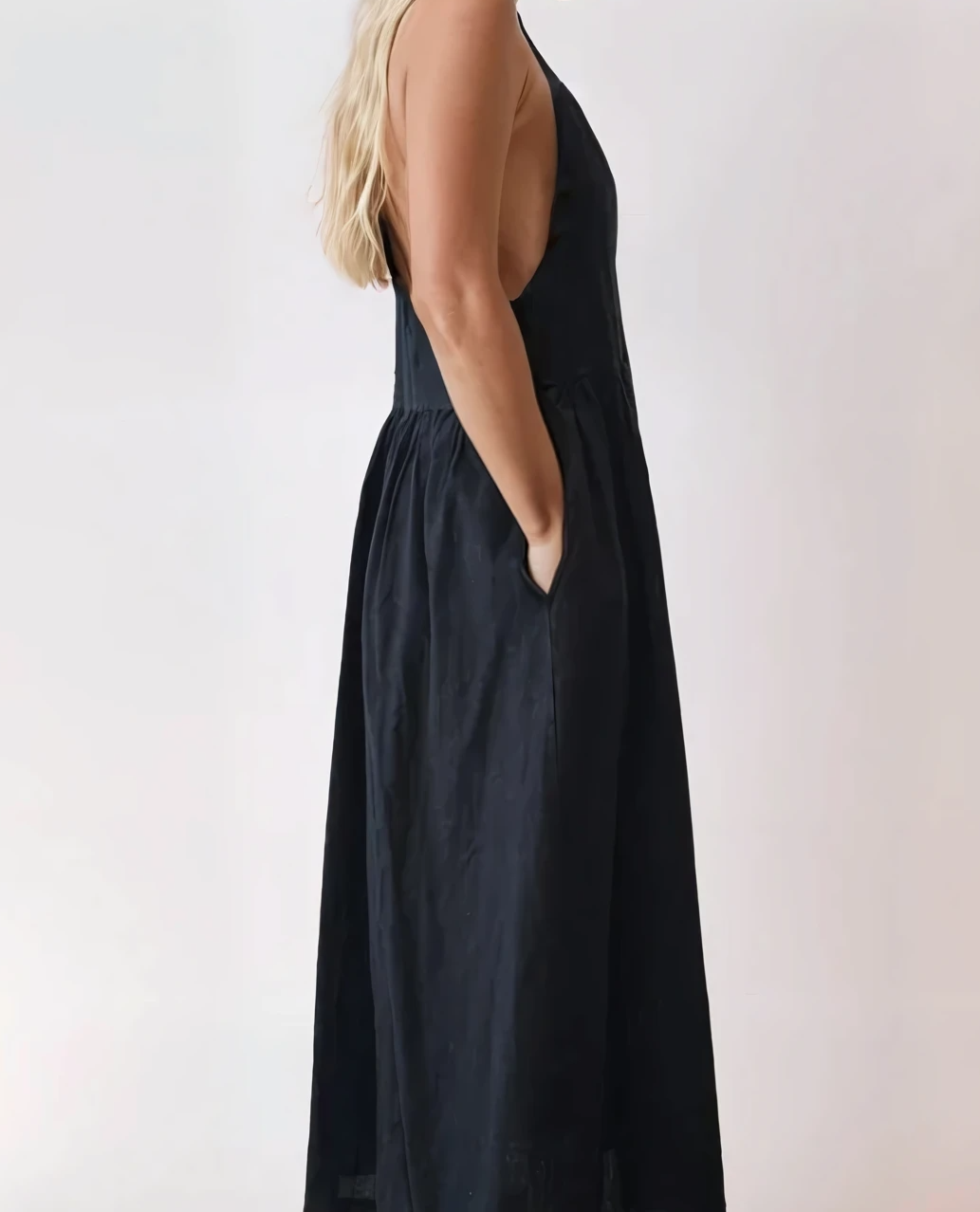 Side profile of black pinafore dress showing open sides, deep armholes, and relaxed fit with hands in pockets.