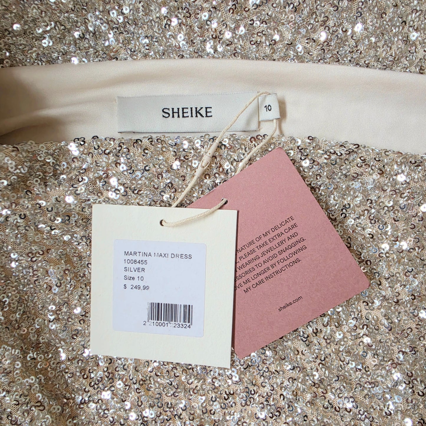 A close-up of the Martina sequined dress with a visible SHEIKE brand label and care tag. The garment is covered in silver glitter sequins.
