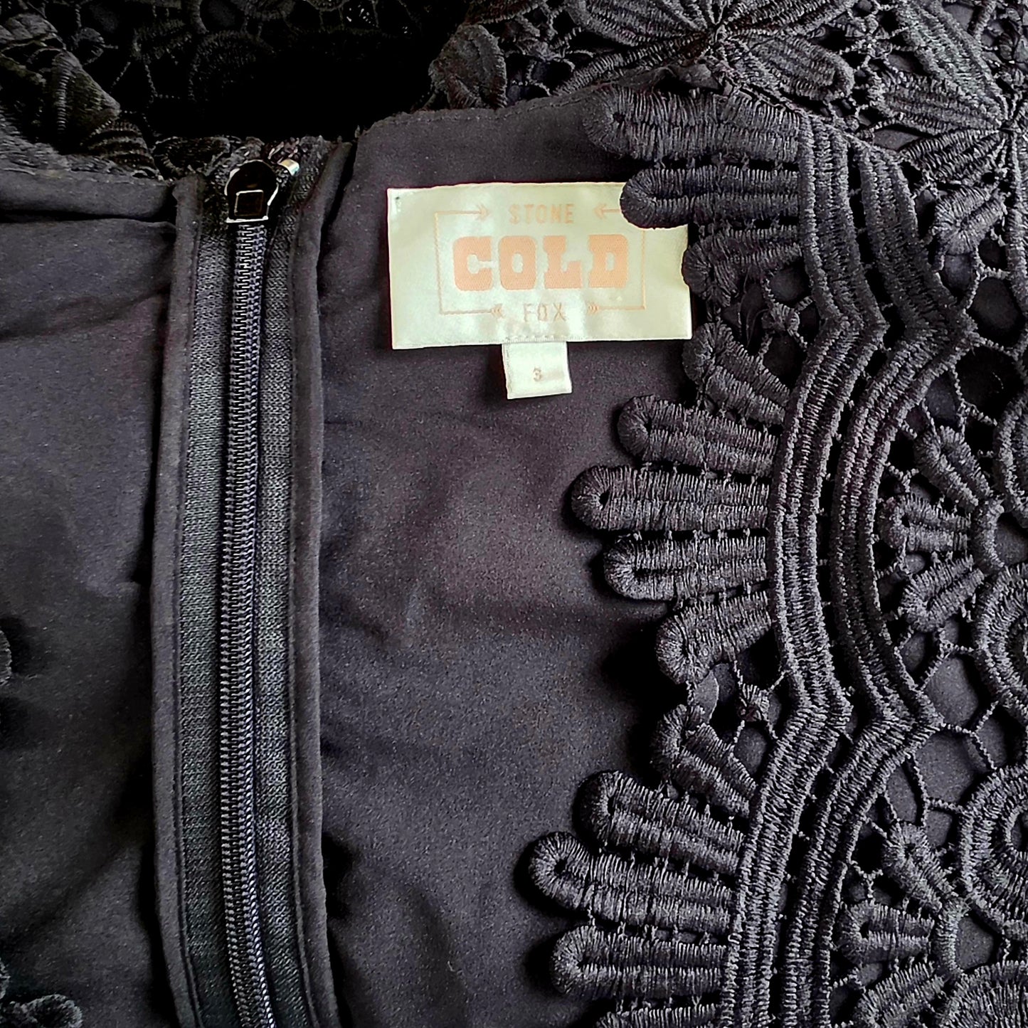 Close up view of the Stone Cold Fox Saint Jumpsuit's neckline, showing the lace, zip up back and brand label.