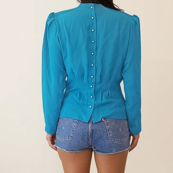 Model shown from behind wearing vintage teal pleated blouse with structured fit and full-length sleeves, featuring mini shoulder pads and elegant button closure.