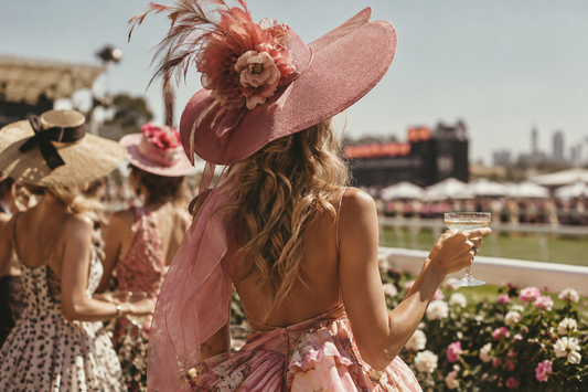 Woman wearing an oversized pink floral hat with feathers and a floral dress at a sunny race-day event, holding a champagne glass.