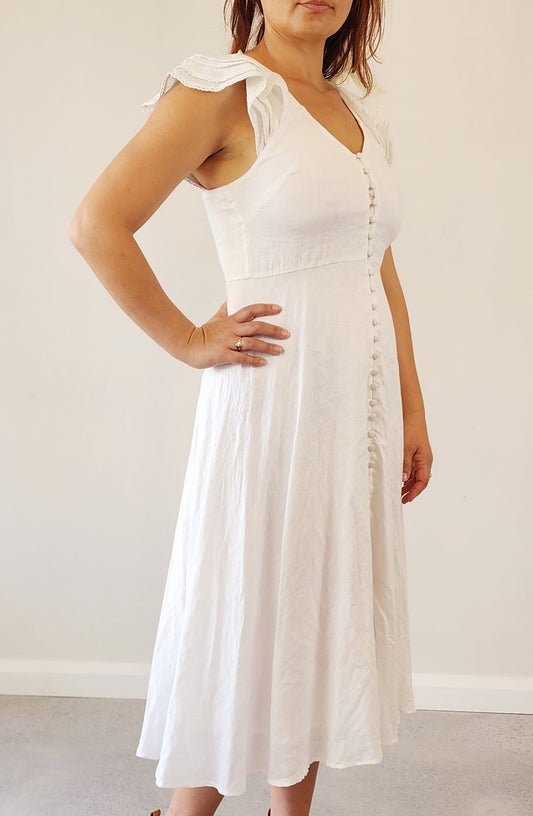 Front view of a woman wearing the Bec & Bridge Lafontelina dress — a crisp white linen midi with flutter sleeves and fabric-covered buttons down the bodice.