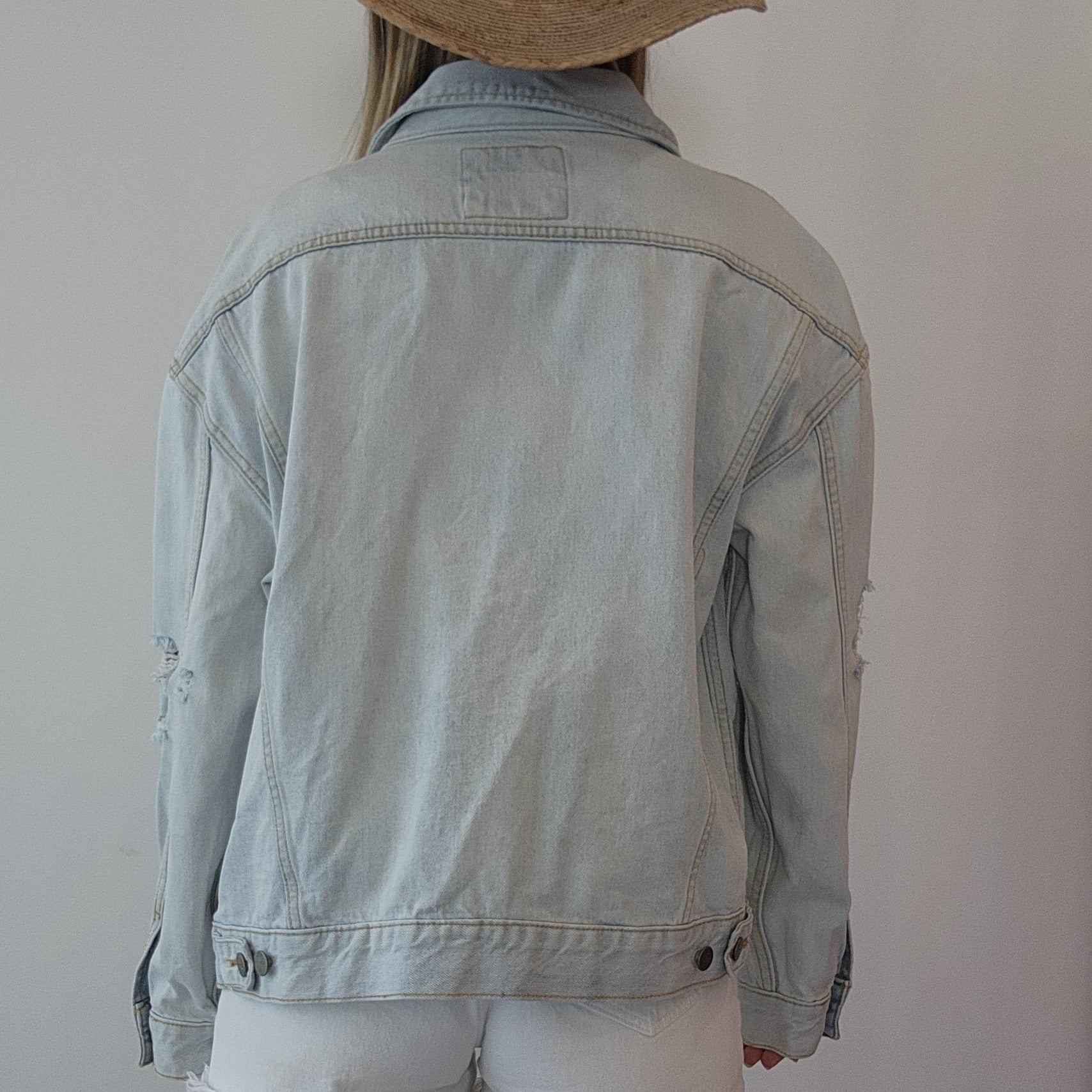 Back view of a model wearing a light wash denim button up jacket by clothing designer RVCA. Distressed style, made to last.