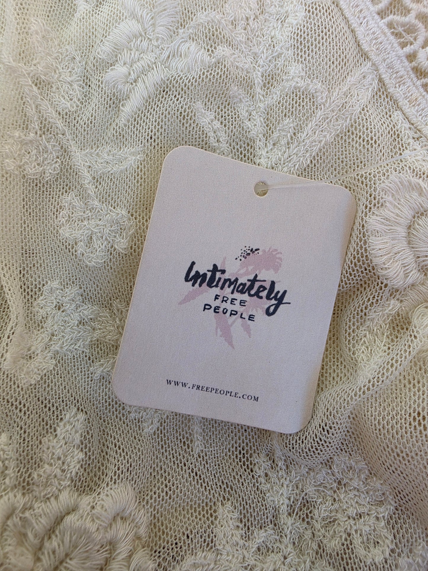 A close-up of a cream-colored lace dress with a paper hangtag reading 'Intimately Free People' and the website www.freepeople.com
