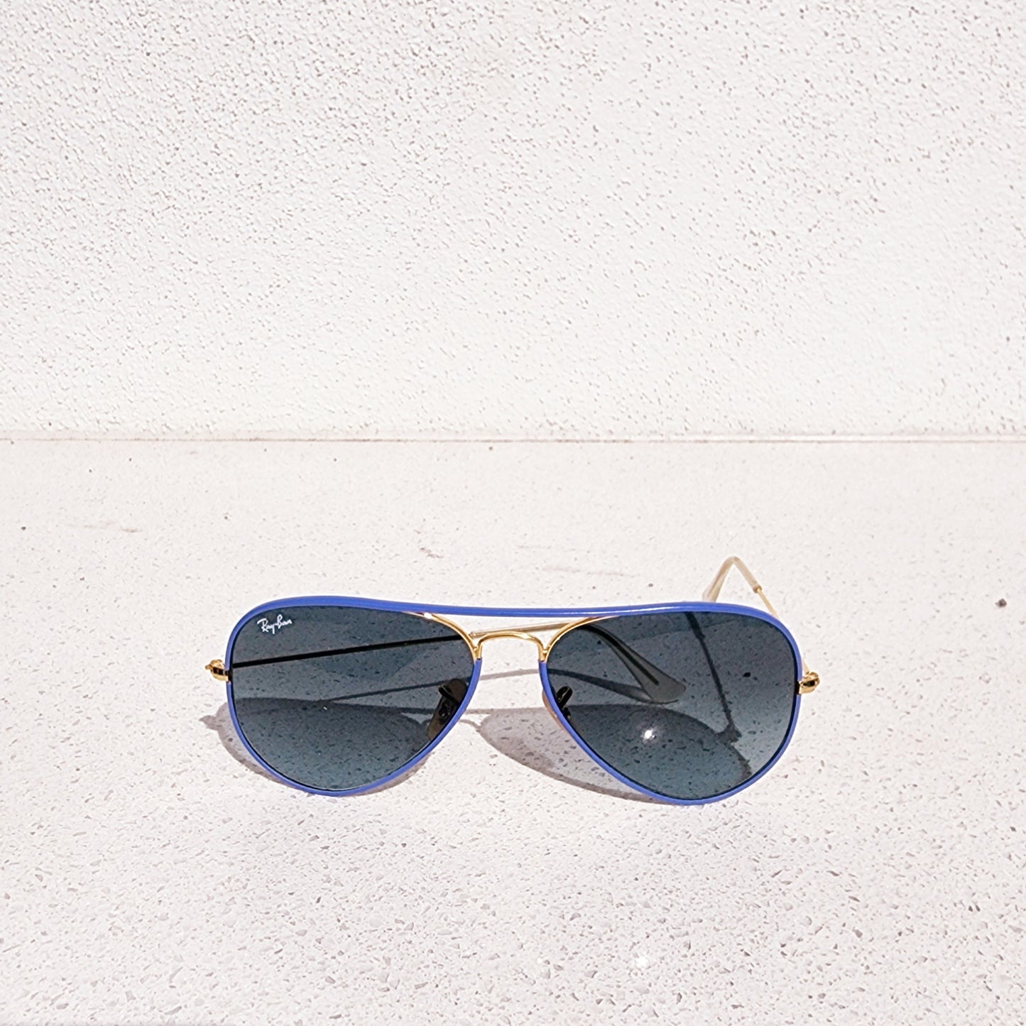Ray Ban full colour aviator sunglasses with blue lenses and gold frame on a white textured surface.
