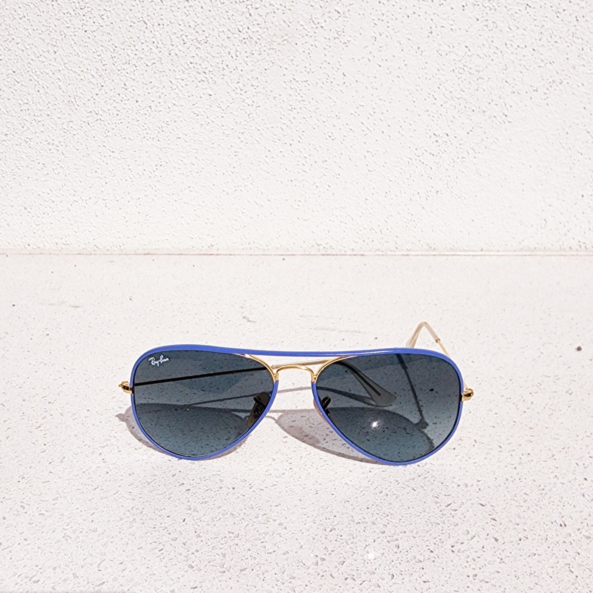 Ray Ban full colour aviator sunglasses with blue lenses and gold frame on a white textured surface.