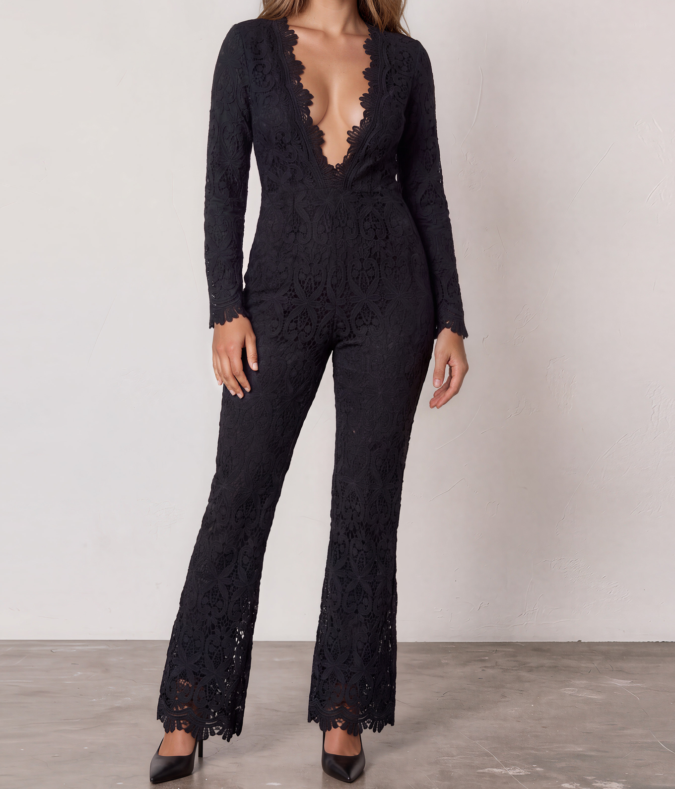 A woman modeling the Stone Cold Fox black lace long-sleeve Saint jumpsuit with a deep V-neckline and slightly flared legs