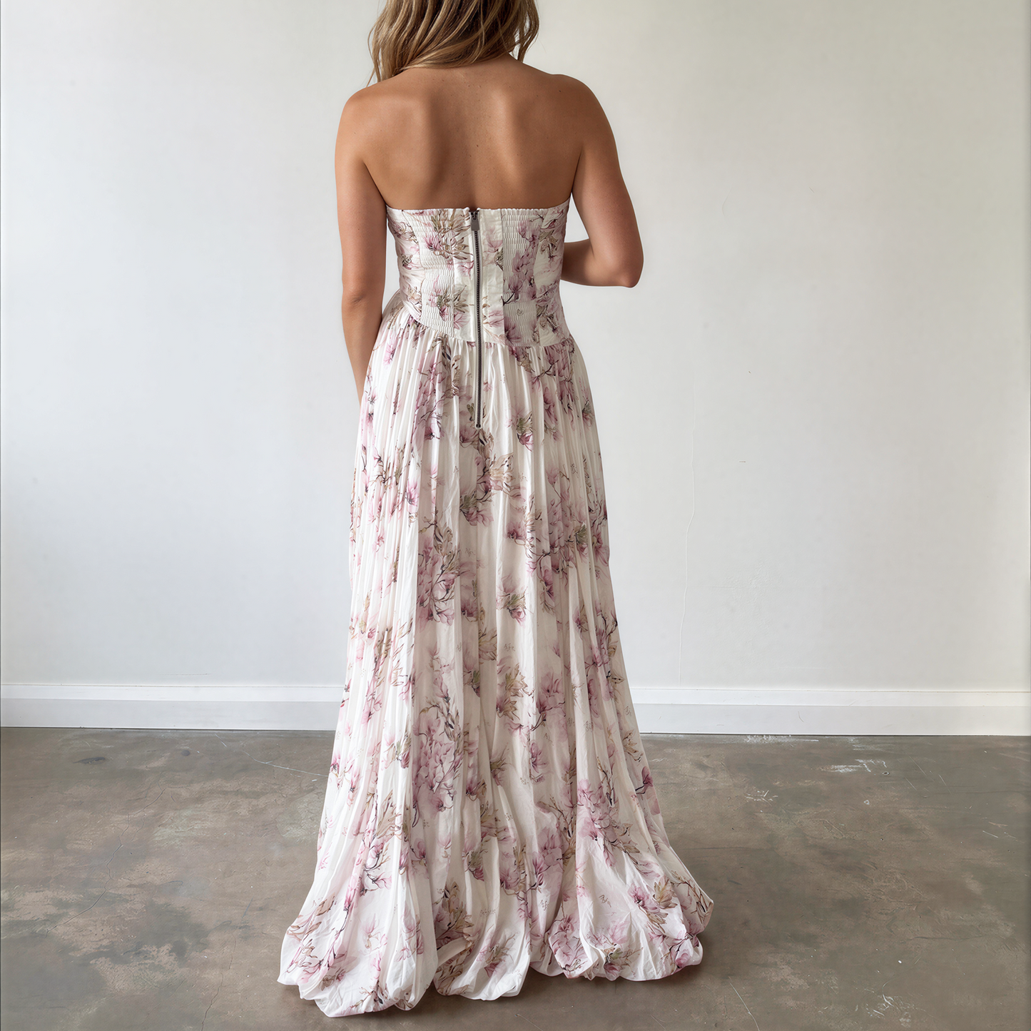 Back view of a model wearing the Aje strapless white maxi dress with a pink floral print showing the zip up back and bubble hemline.