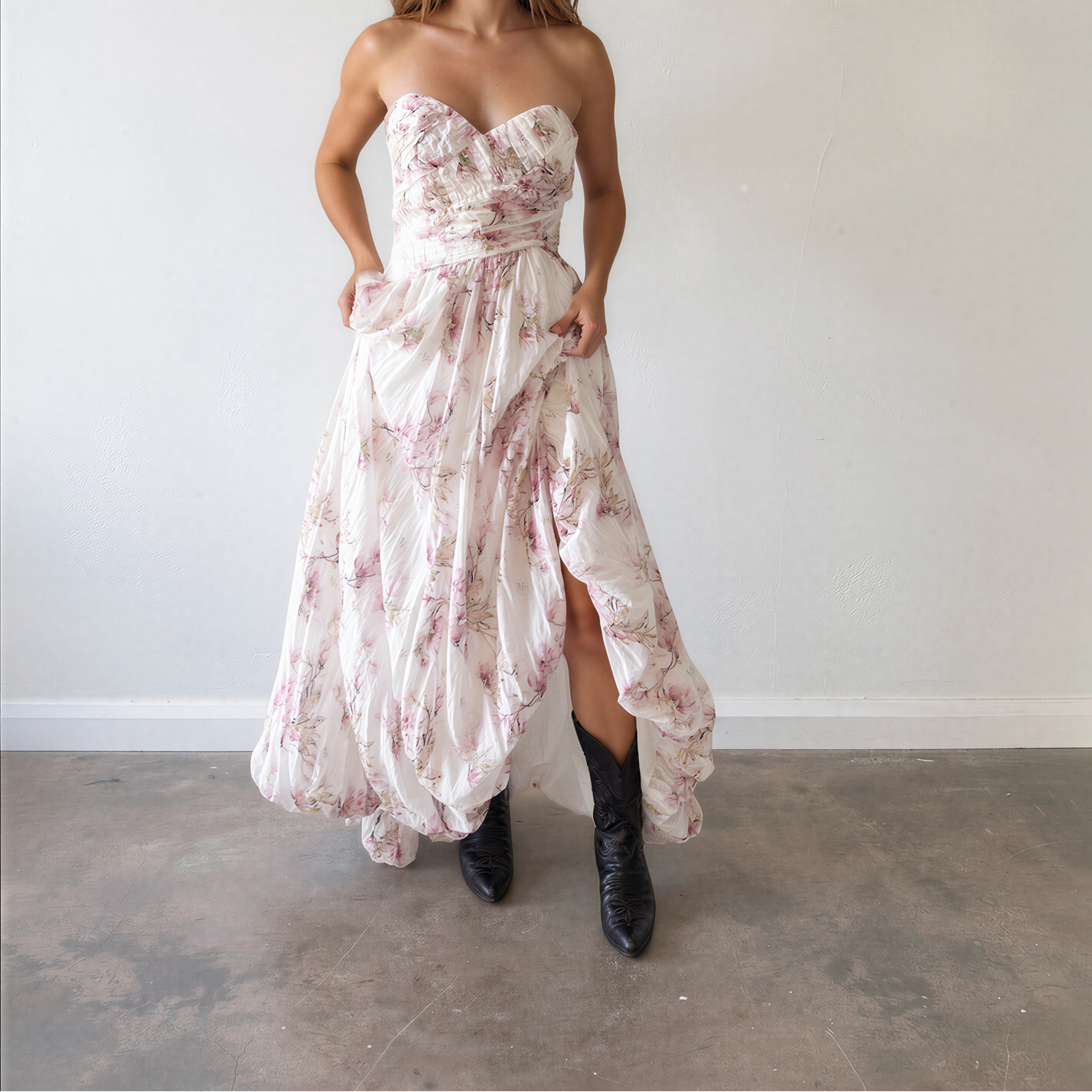Front view of a model wearing the Aje strapless white maxi dress with a pink floral print and a high-low ruffled hem, paired with black cowboy boots