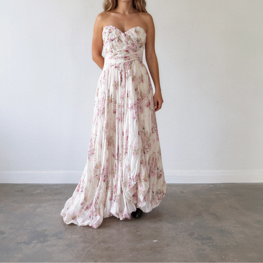 A woman showing the front view of the Aje strapless maxi dress with a sweetheart neckline, a fitted waist, and a flowing skirt with a pink floral print on a cream background.