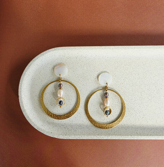 A styled image of the Auricle pearl hoop earrings, showing the large brass hoop, multi-coloured pearls and mother of pearl elements.