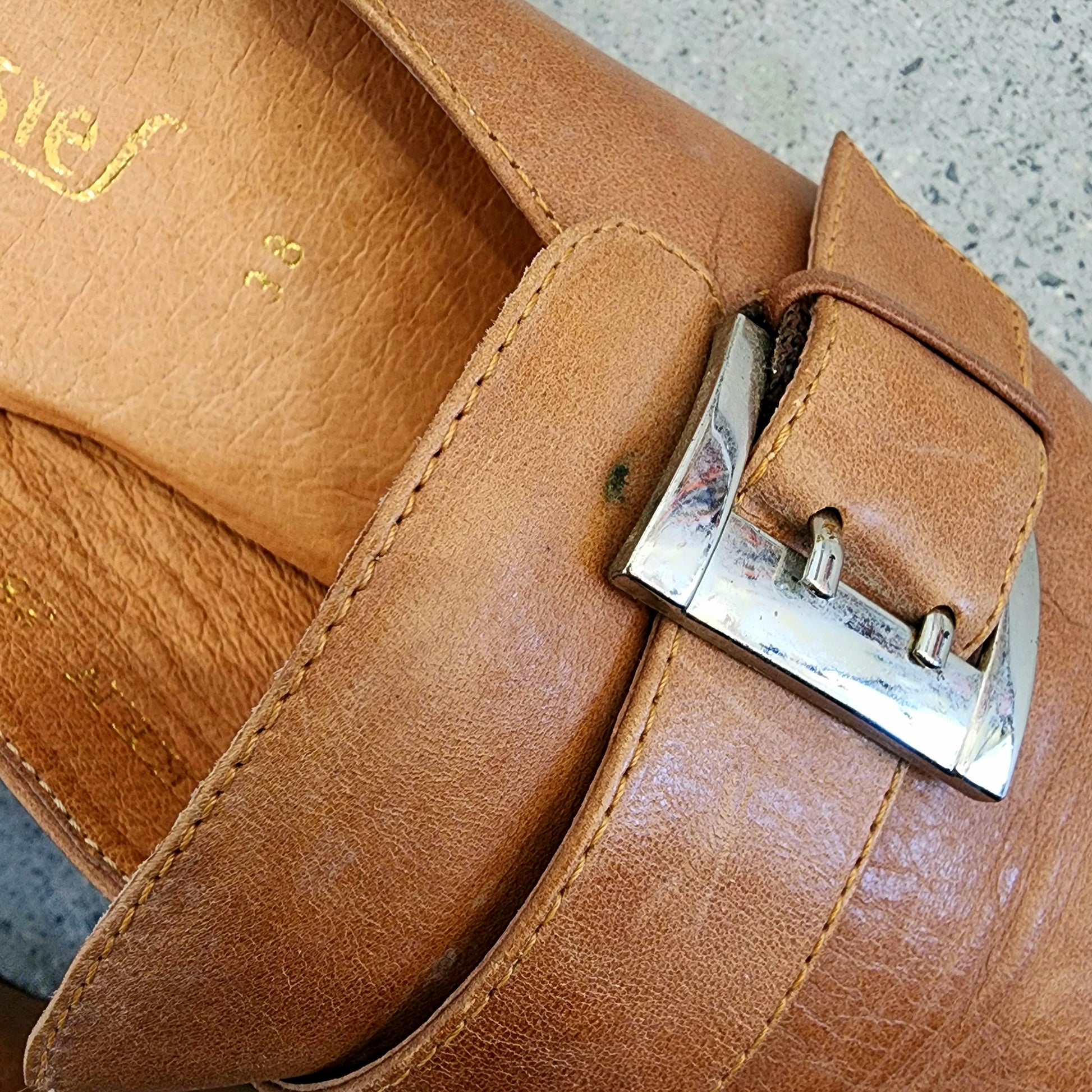Close-up of the Chrissie tan mule buckle, showing the metal hardware and leather strap detail.