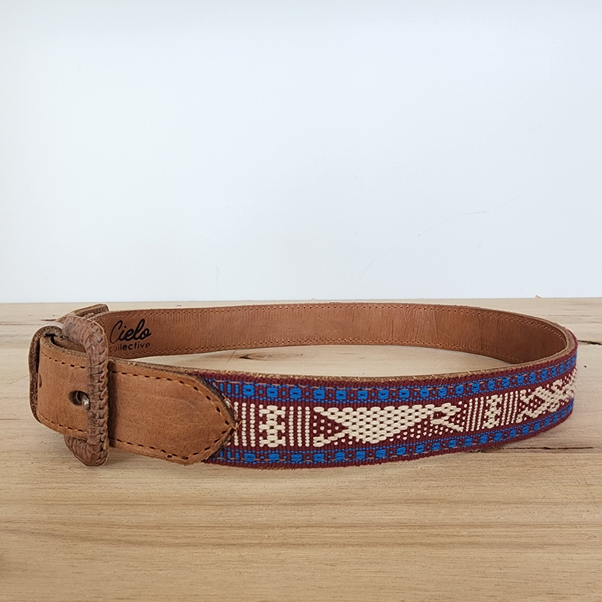 Handwoven Cielo Collective belt with colourful woven panel and tan leather strap displayed on wooden surface.