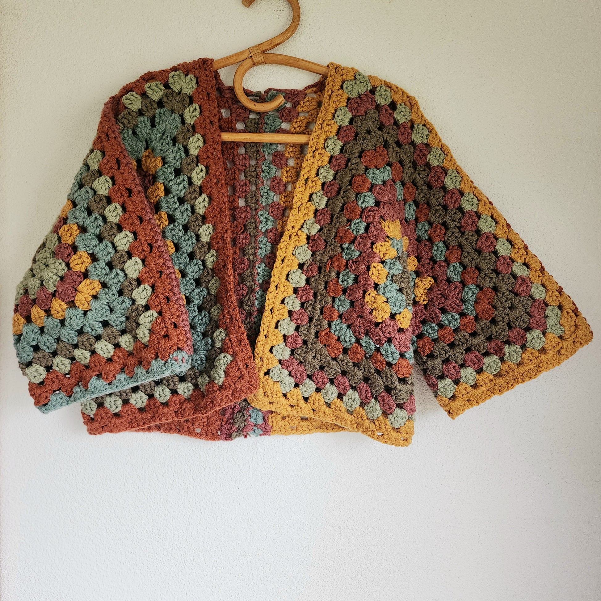 Hand-crocheted granny square cardigan in earthy mustard, sage and rust tones hanging on wooden hanger.