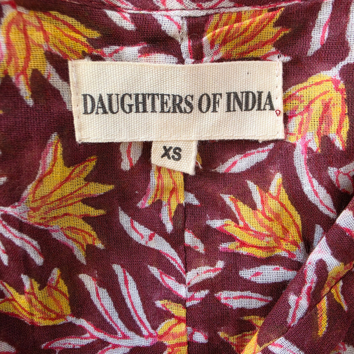 Close-up of a patterned fabric garment with a visible sewn-in label reading DAUGHTERS OF INDIA and a size tag reading XS.