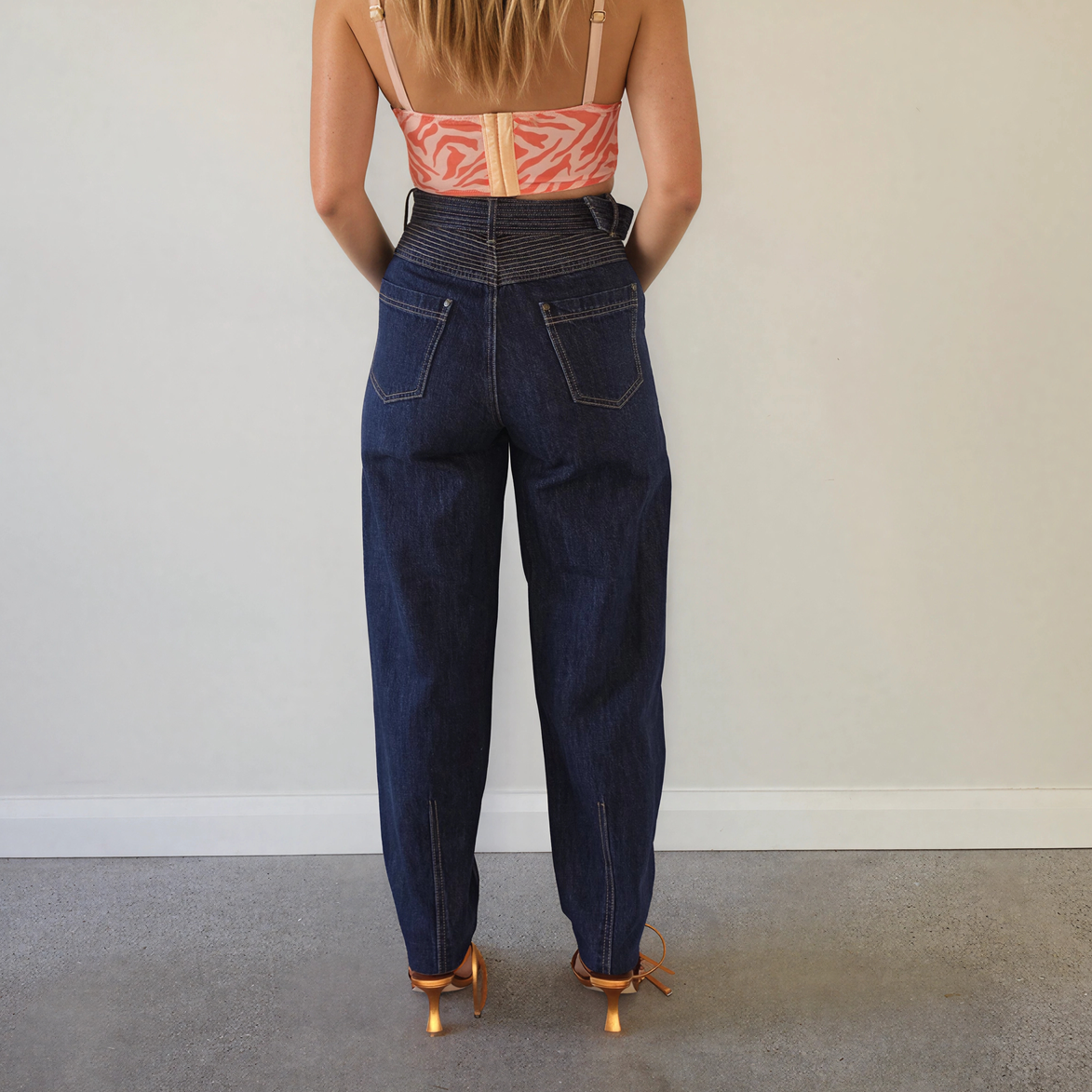  The back view of a woman wearing the Kivari Adina Jeans in Indigo, showing the high waist, pocket details, belted style and tapered leg.