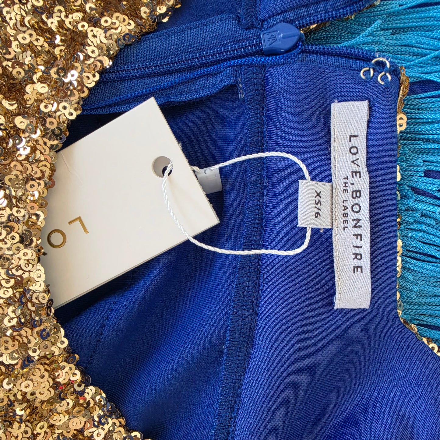 Close-up of the Love Bonfire Jewel Skies fringe dress, showing the gold sequined edge, turquoise fringe trim, and visible tags reading LOVE BONEFIRE and XS/6.