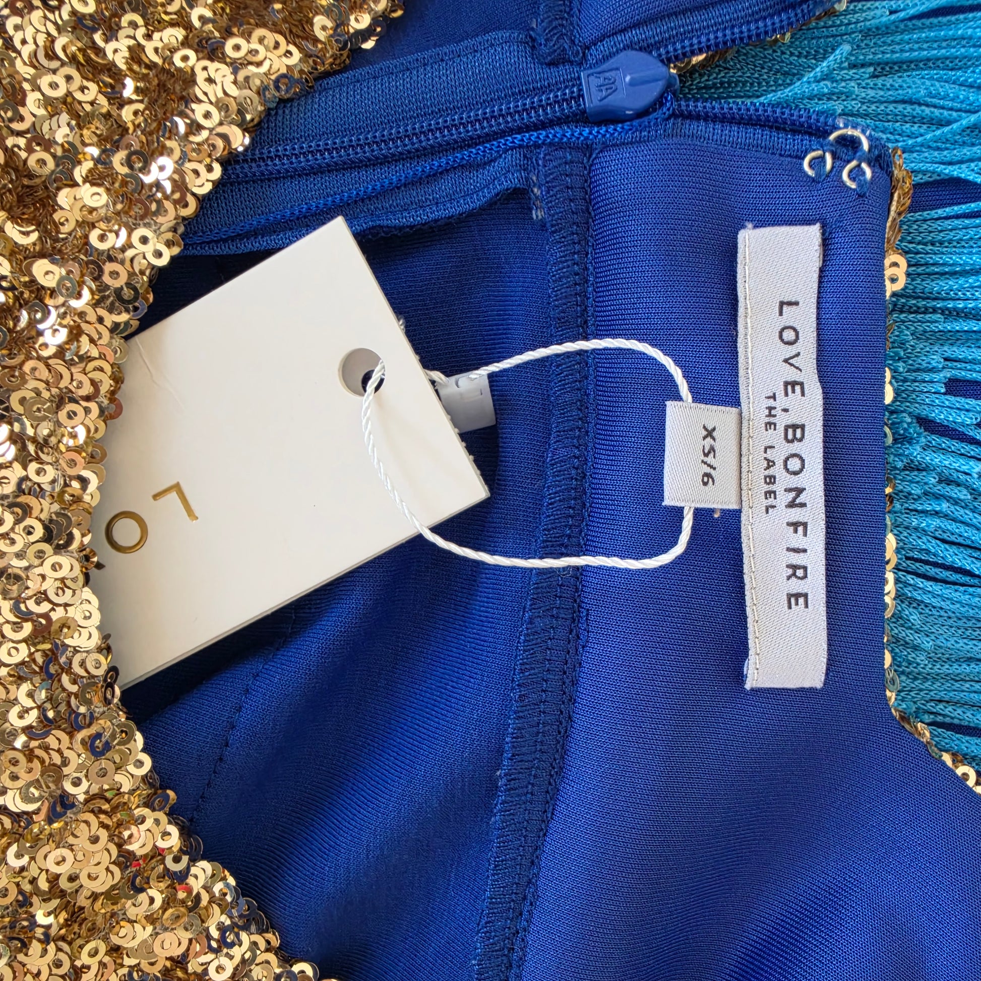 Close-up of the Love Bonfire Jewel Skies fringe dress, showing the gold sequined edge, turquoise fringe trim, and visible tags reading LOVE BONEFIRE and XS/6.