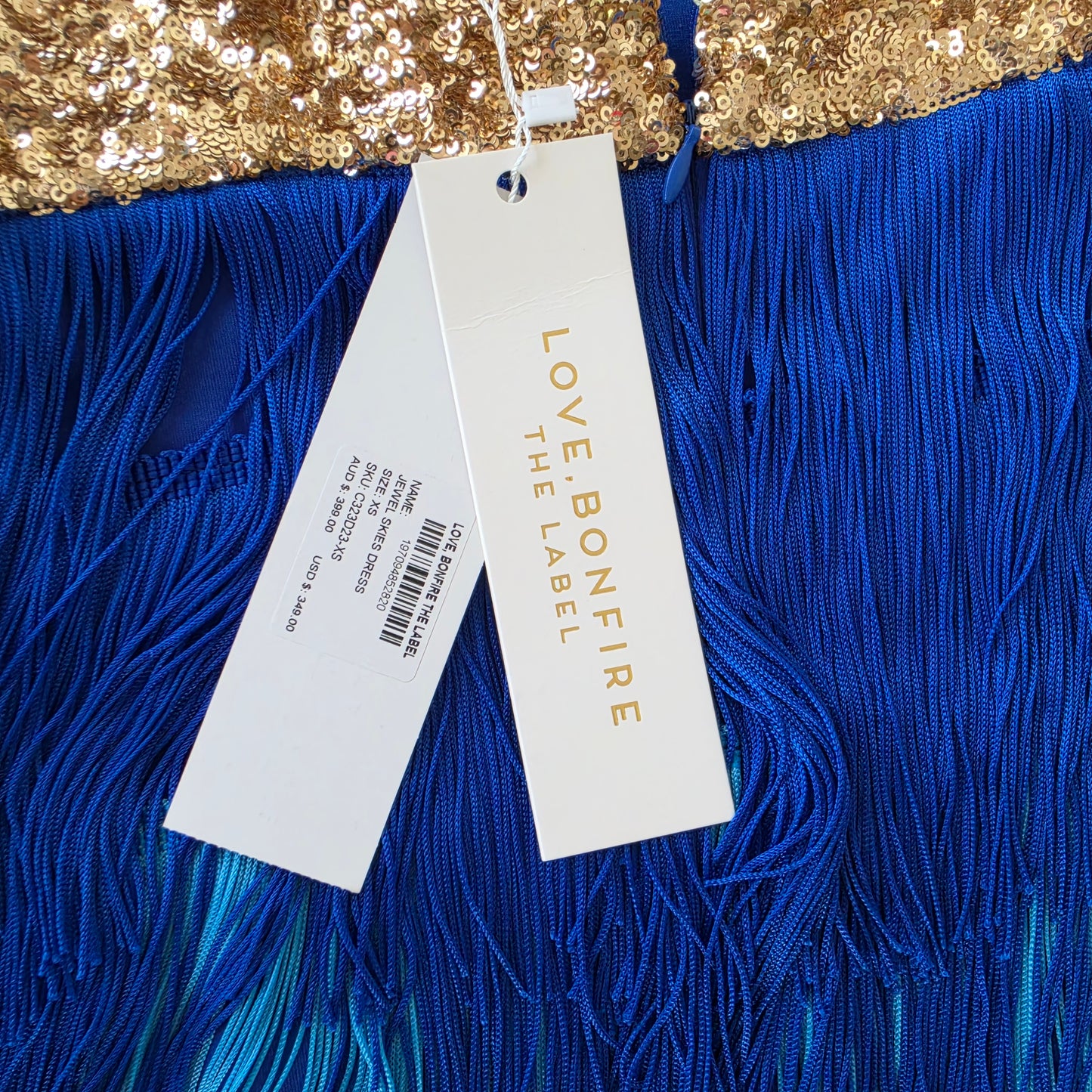 Close-up of the Love Bonfire Jewel Skies blue fringed dress showing the  wide gold sequined trim neckline, blue fringe and garment tags