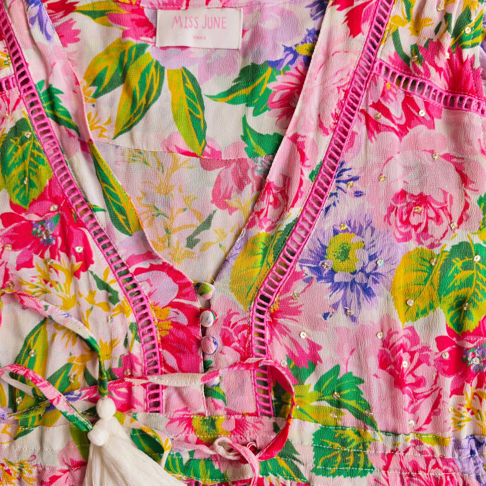 Close-up of a Miss June Paris floral dress featuring a vibrant pink, green, and yellow floral print, with pink crochet trim along the V-neckline, sequin embellished details and waist ties.