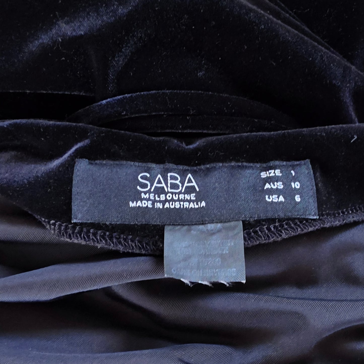 Close-up of a black velvet garment label reading SABA Melbourne Made in Australia, with size information printed on a dark fabric background.