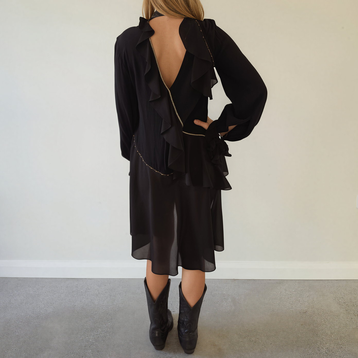Back view of a woman the Sass & Bide Made for Love top, long-sleeve with dramatic ruffle trim, a deep open back, and a thin gold chain detail, paired with black cowboy boots.