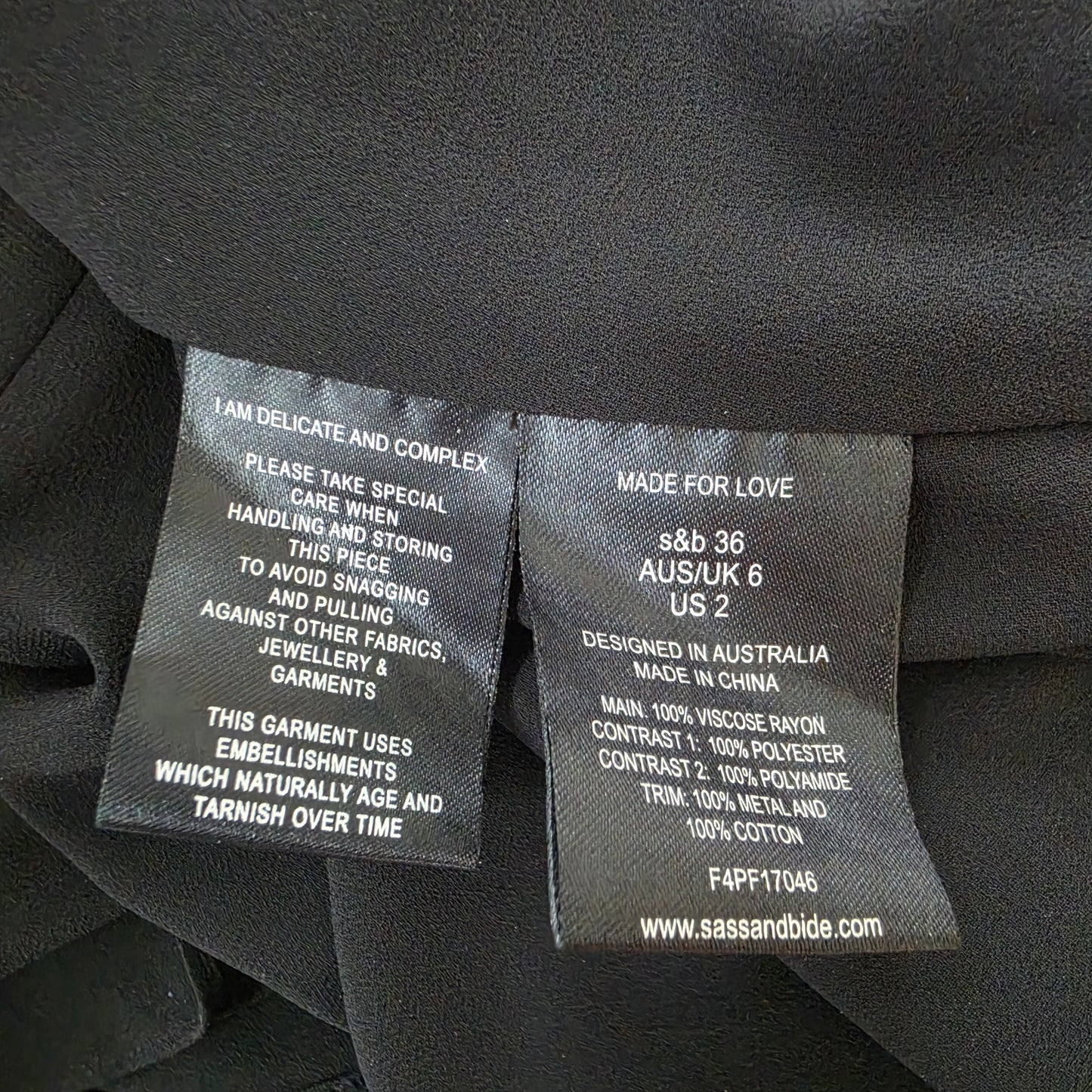 Close-up of black garment labels sewn onto dark fabric, showing text including 'MADE FOR LOVE', 'AUS/UK 6 US 2', and 'www.sissholding.