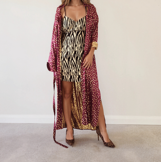 Front view of the Scotch & Soda Magnifique long satin duster dress in burgundy with metallic gold polka dots, tie belt and side splits, worn with a metallic zebra dress and leopard print heels.