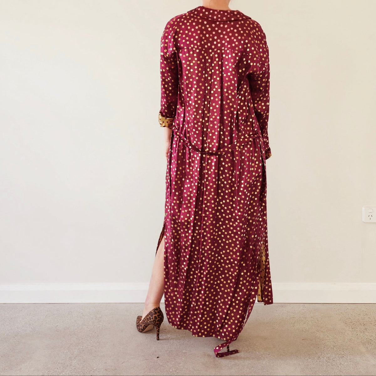 Back view of Scotch & Soda Magnifique long satin duster in burgundy with metallic gold polka dots and self-tie belt, worn with leopard print heels.
