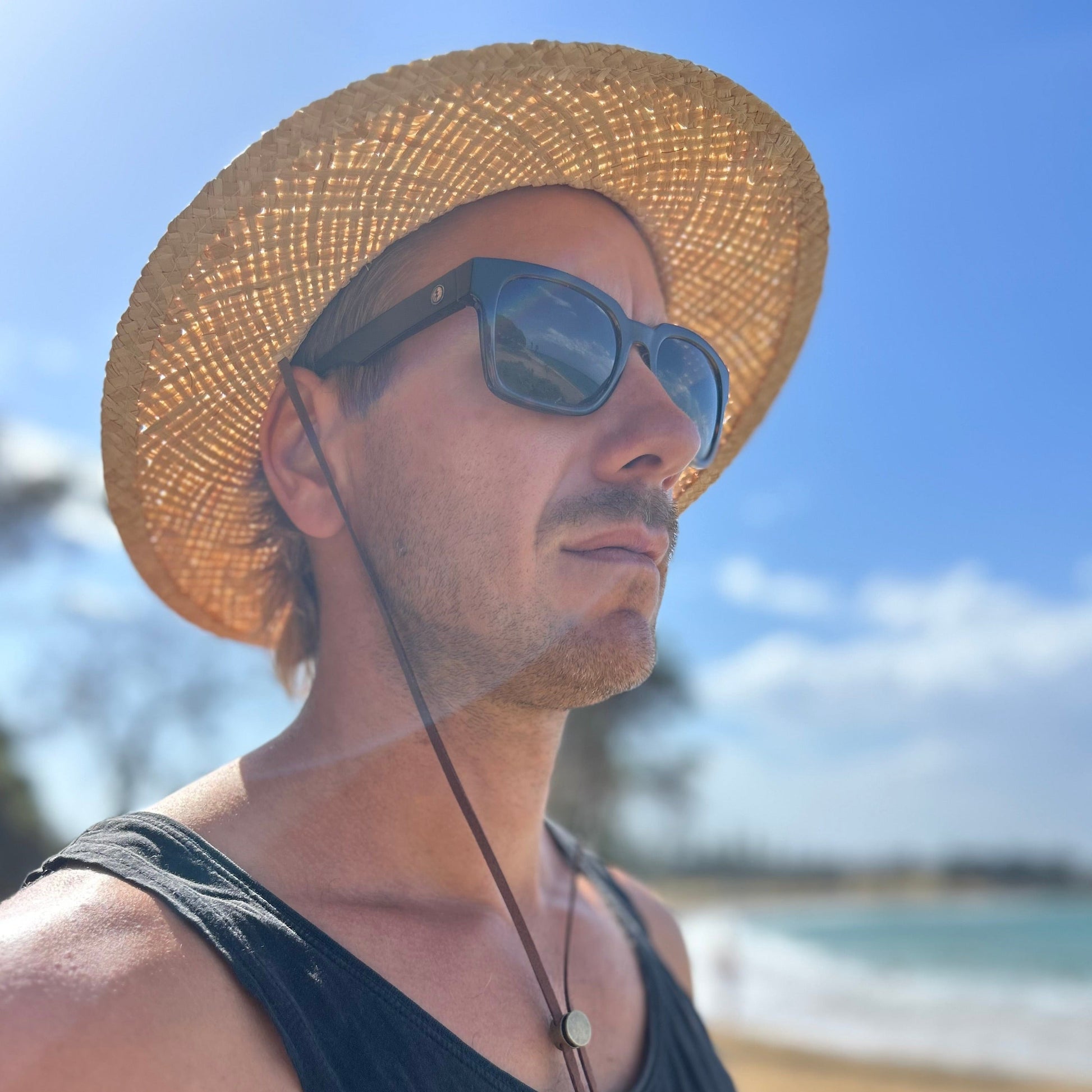 Man wearing a Shayd straw hat and sunglasses with a beach in the background