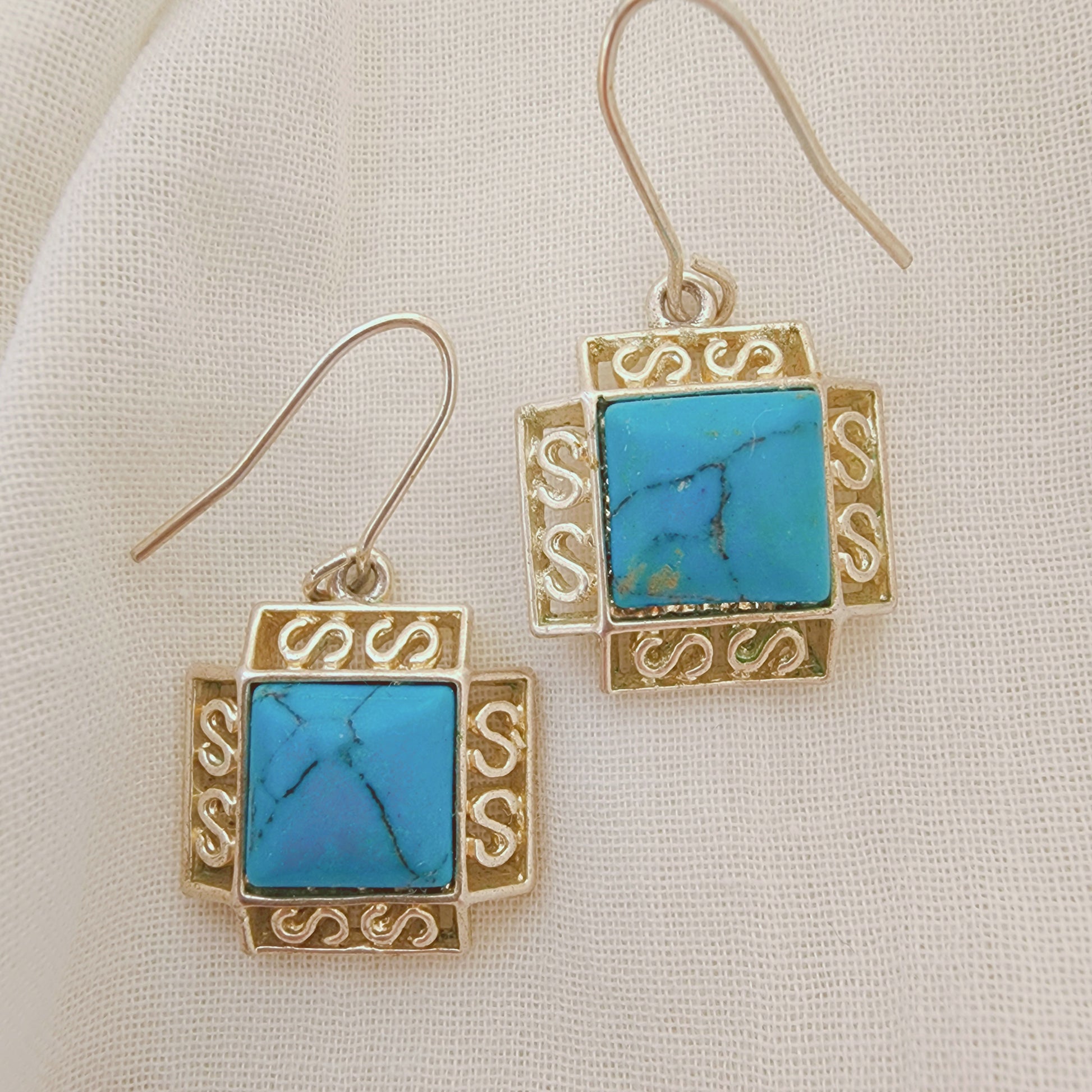 The front view of sterling silver earrings with turquoise-coloured stones in a square setting, photographed on soft neutral fabric.