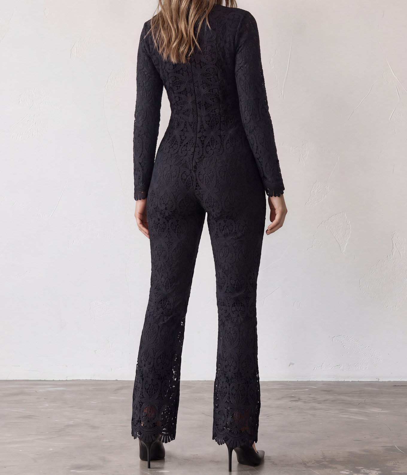 The back view of a womane wearing the Stone Cold Fox black lace long-sleeve Saint jumpsuit with a high back zip and slightly flared legs