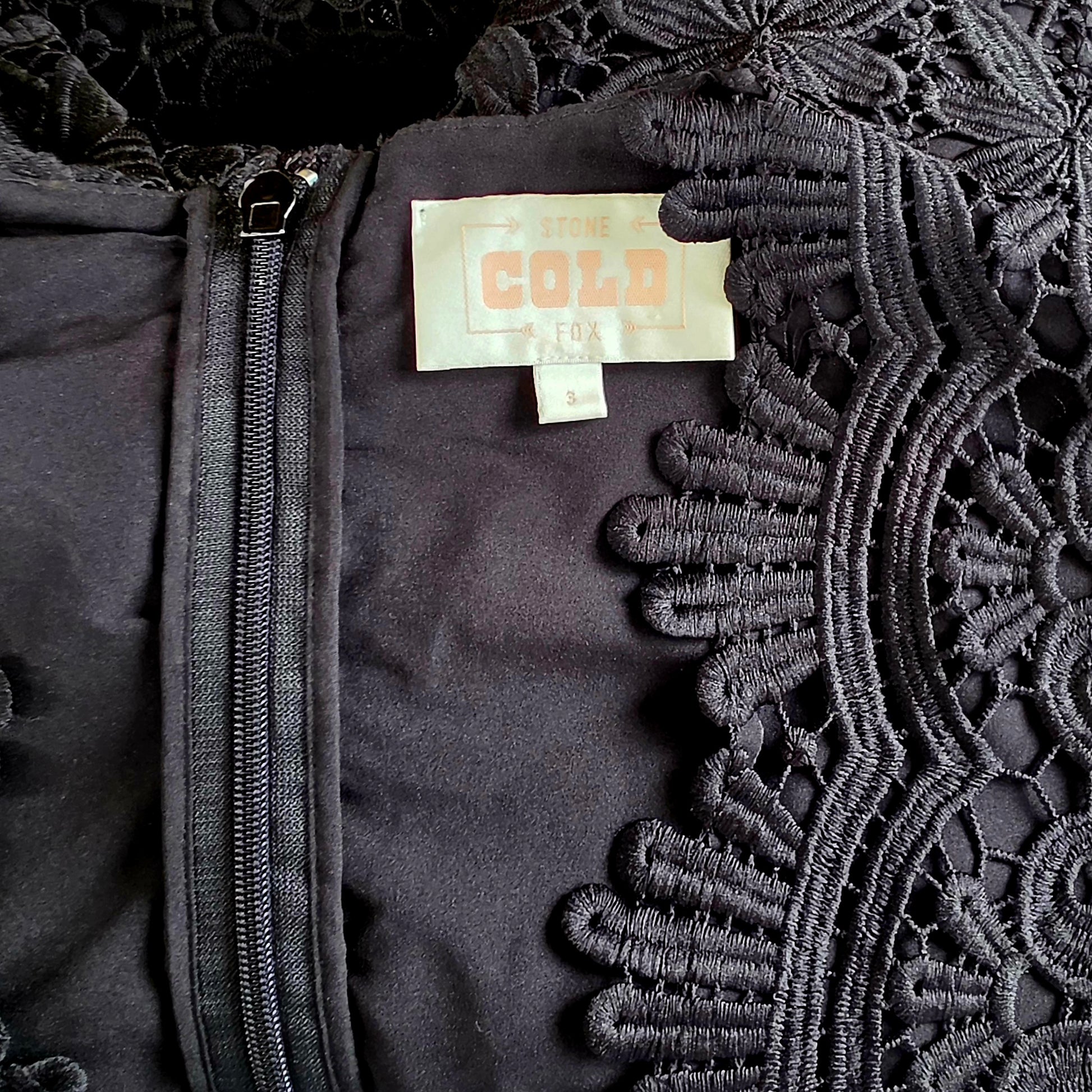 Close up view of the Stone Cold Fox Saint Jumpsuit's neckline, showing the lace, zip up back and brand label.