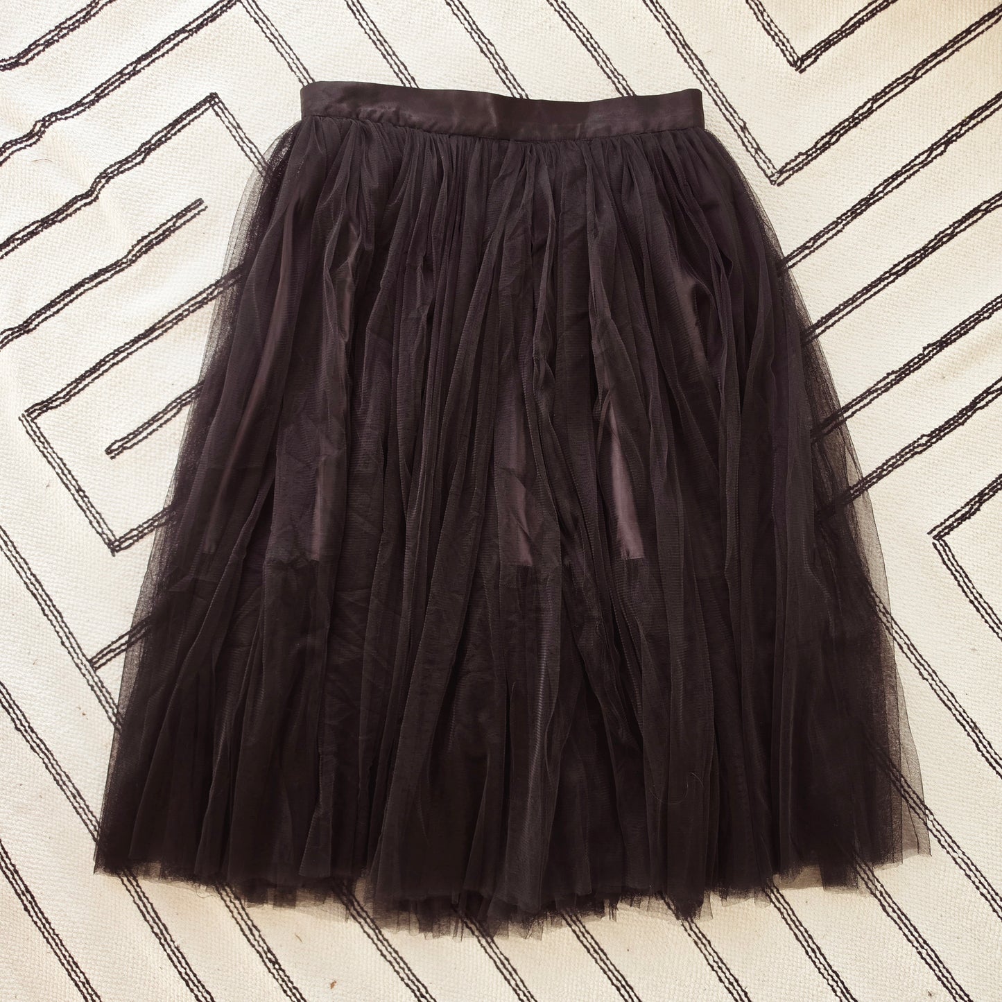 A black Style Workroom tulle tutu skirt laid flat on a cream-colored rug with black geometric line patterns.