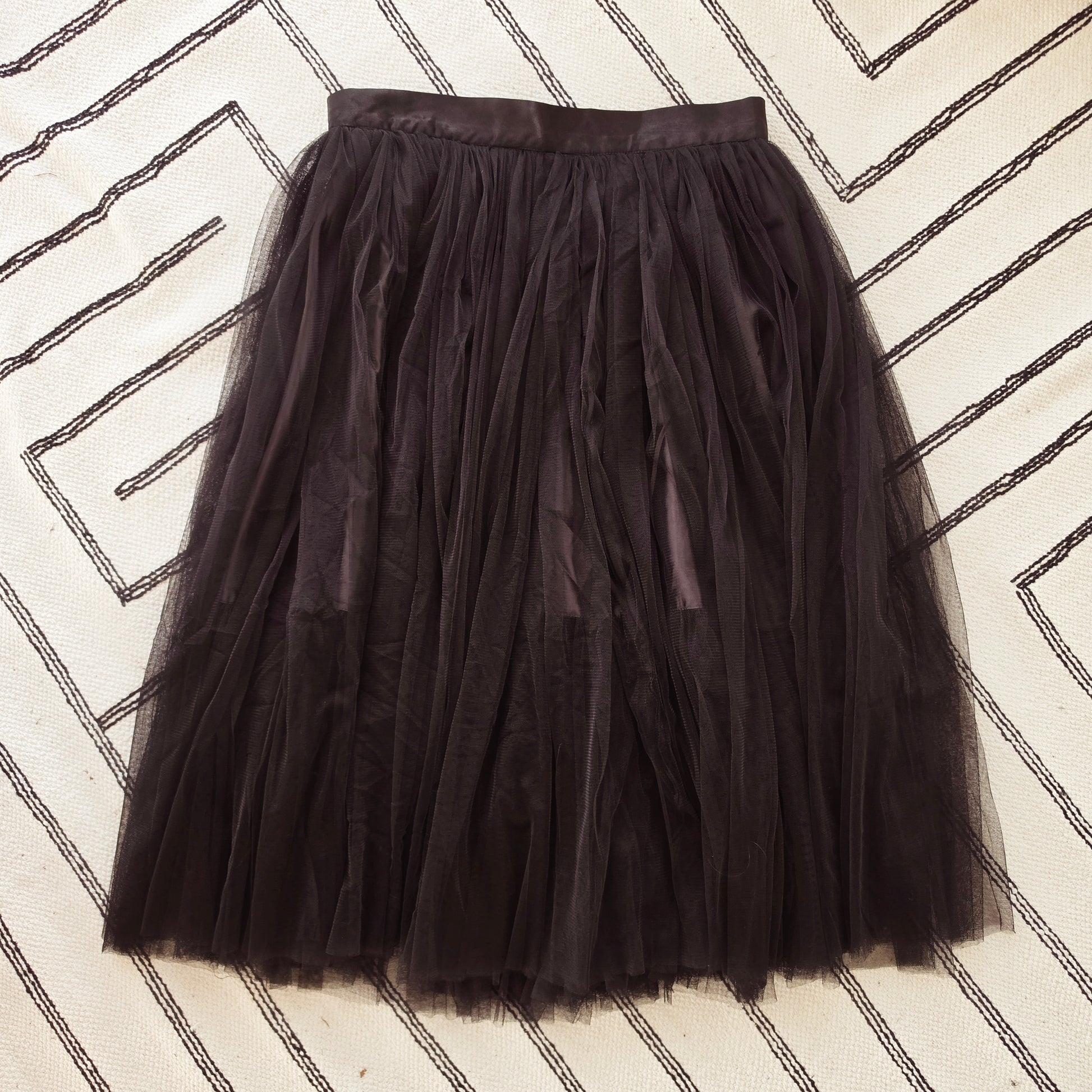 A black Style Workroom tulle tutu skirt laid flat on a cream-colored rug with black geometric line patterns.